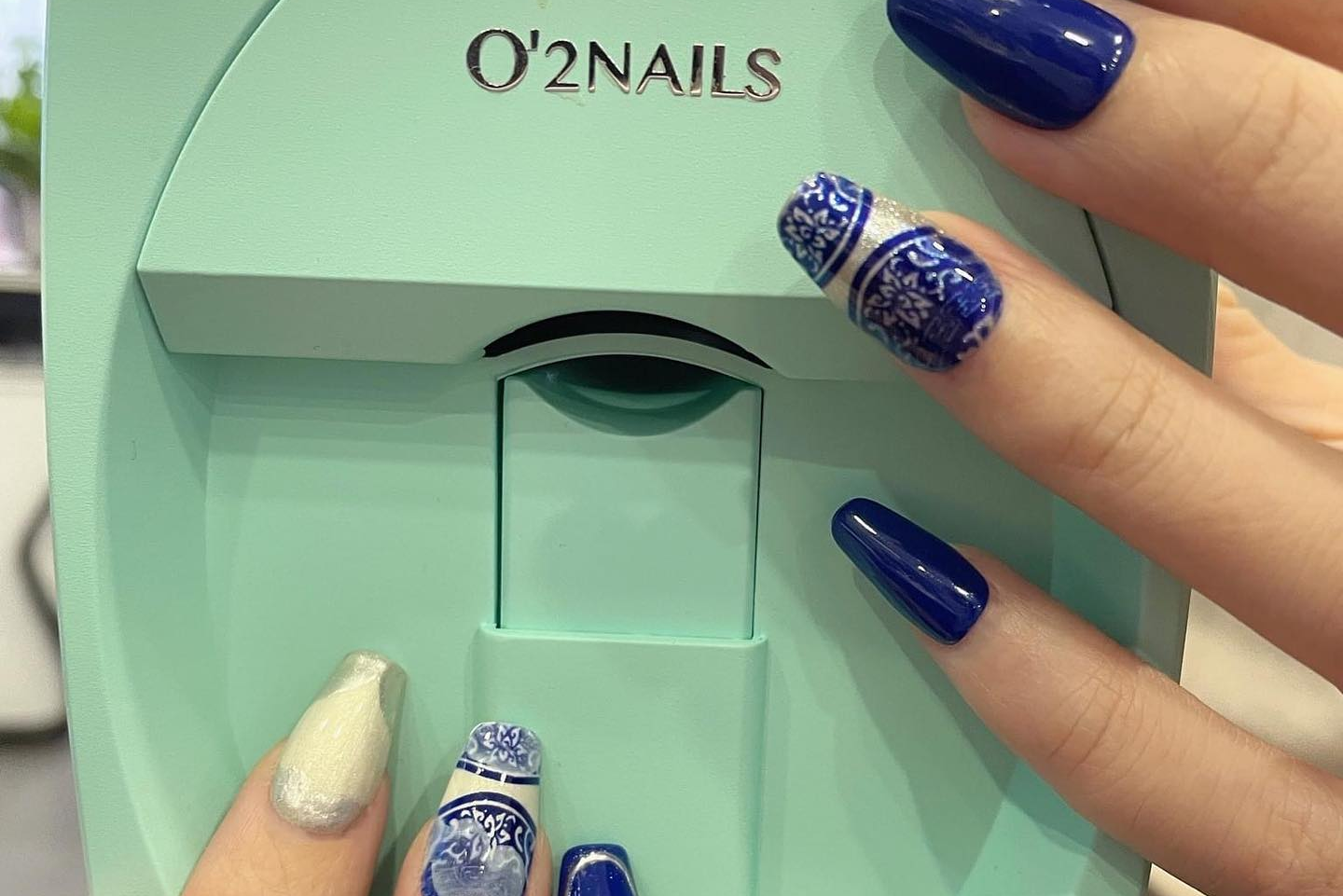 O'2NAILS nail polish machine with hands displaying blue and silver nail designs.